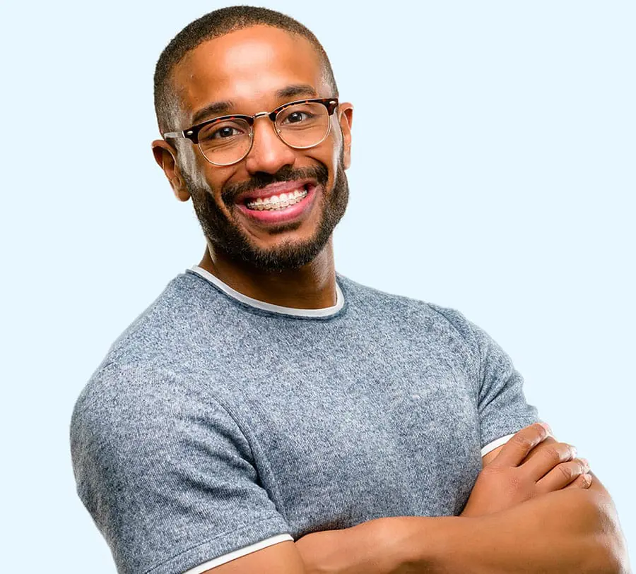 Smiling man with glasses and beard, wearing a gray t-shirt, stands with arms crossed against a light blue background, conveying confidence and warmth.