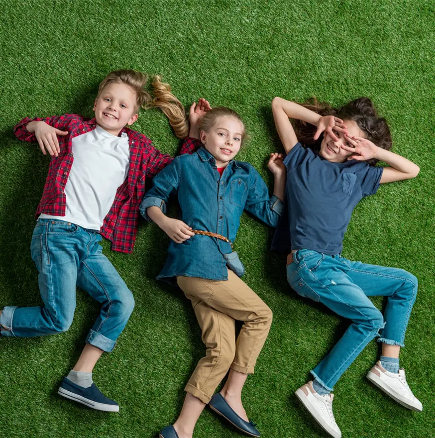 Three children lie on grass, smiling and relaxed. They wear casual clothes: a plaid shirt, denim tops, jeans, and sneakers, conveying a playful, joyful mood.