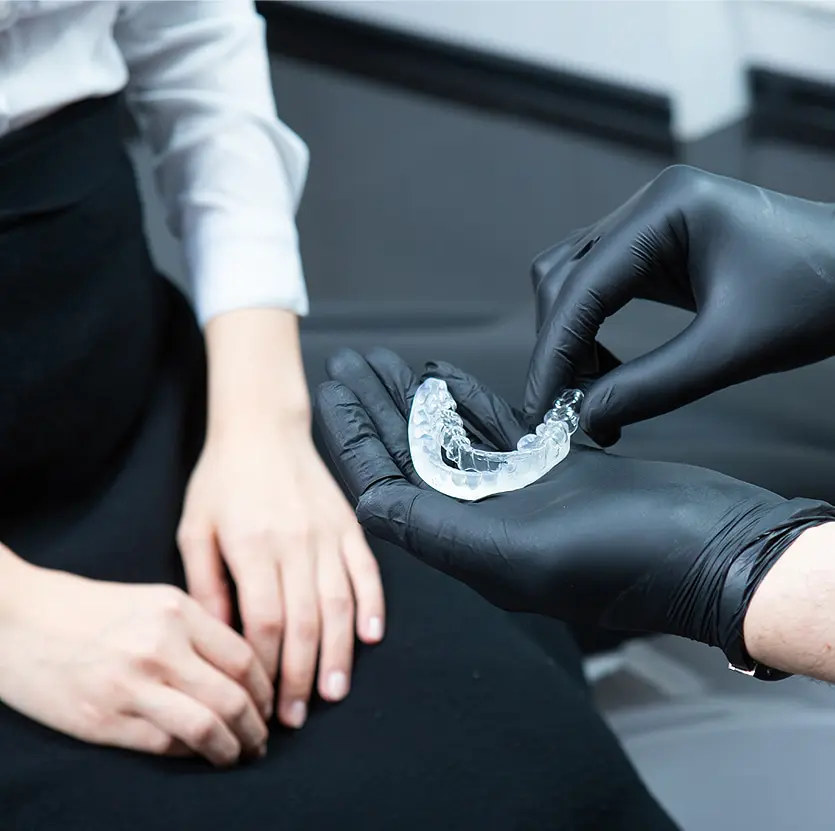 A dentist wearing black gloves holds a clear dental retainer, showing it to a patient in a white blouse and black skirt, seated and attentive.