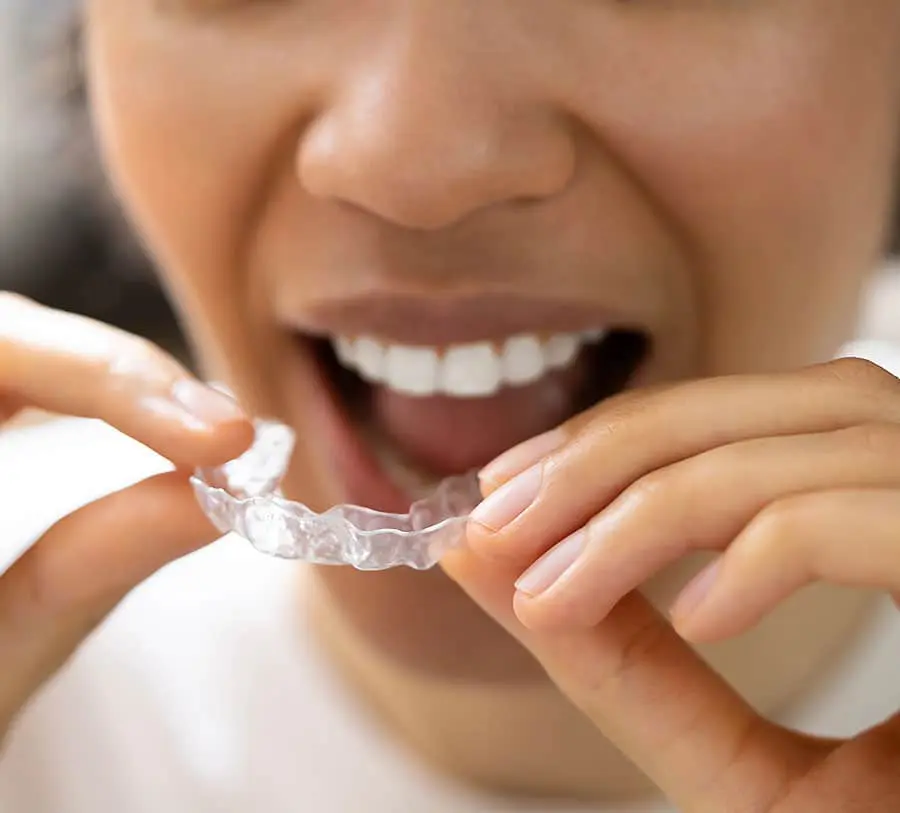 A person smiling while holding a clear dental aligner near their mouth, preparing to insert it. The image conveys a sense of dental care and hygiene.