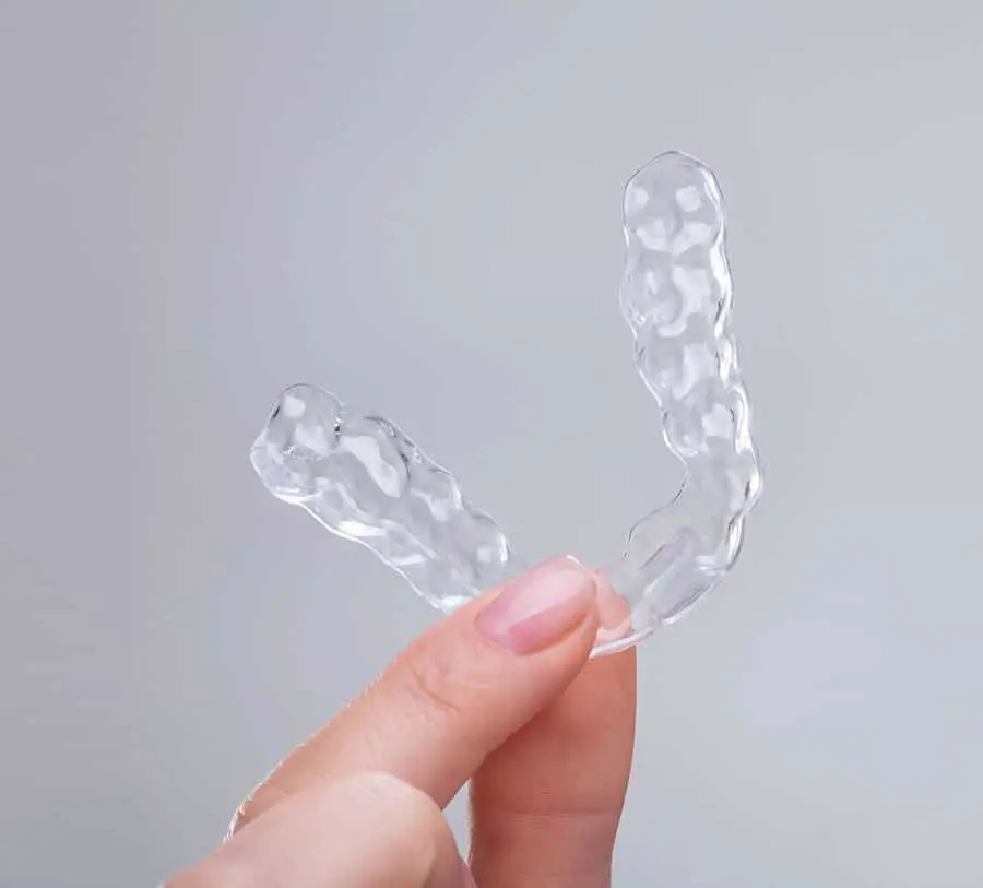 A person holds a clear, textured dental aligner between their thumb and index finger against a plain light gray background.