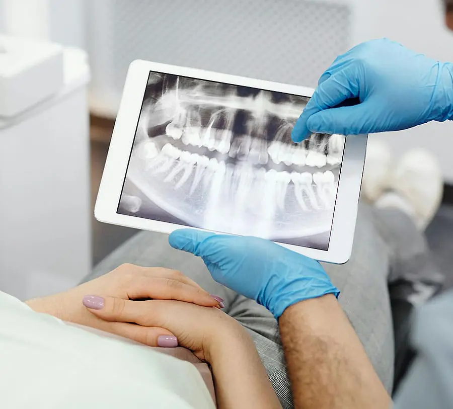 A dentist wearing blue gloves points at a dental X-ray displayed on a tablet. A patient, with folded hands, looks on attentively.
