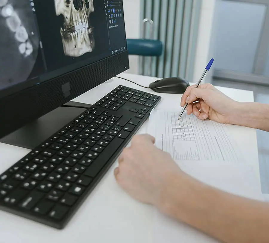 A person writes on a form at a desk, with a computer showing a dental X-ray.