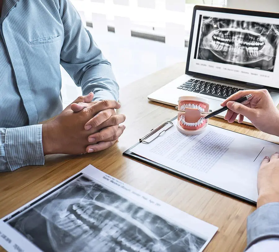 A dentist consults a patient, pointing at a dental model on a desk. X-rays and notes are visible, creating a professional and informative atmosphere.