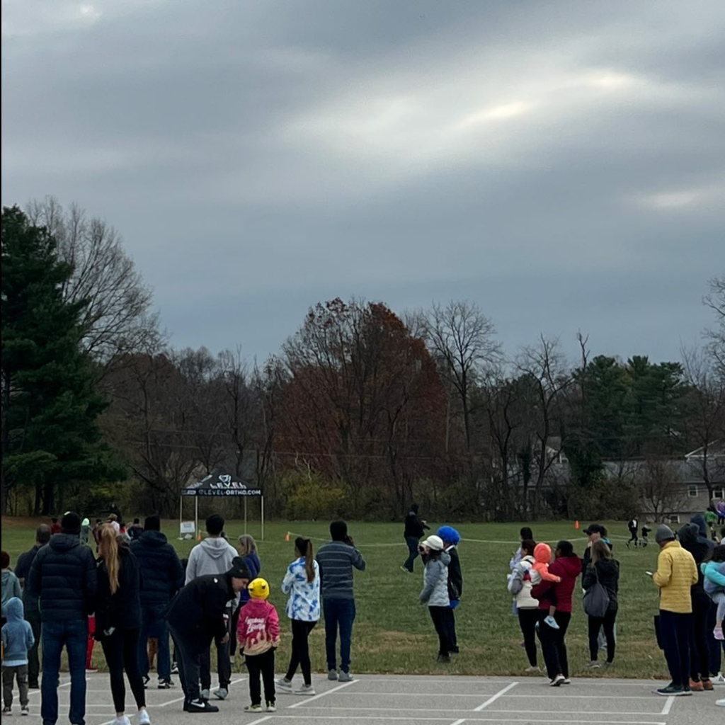 A group of people, including children and adults, stand outdoors on a cloudy day. Some are dressed warmly, and kids wear bright hats, giving a playful vibe.
