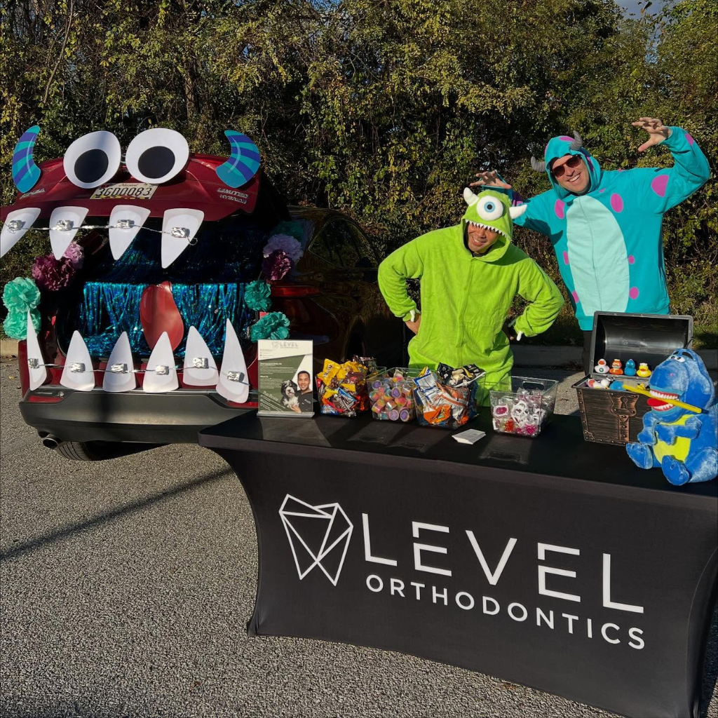 Two people in monster costumes stand next to a car trunk decorated as a monster with teeth and eyes. A table with 